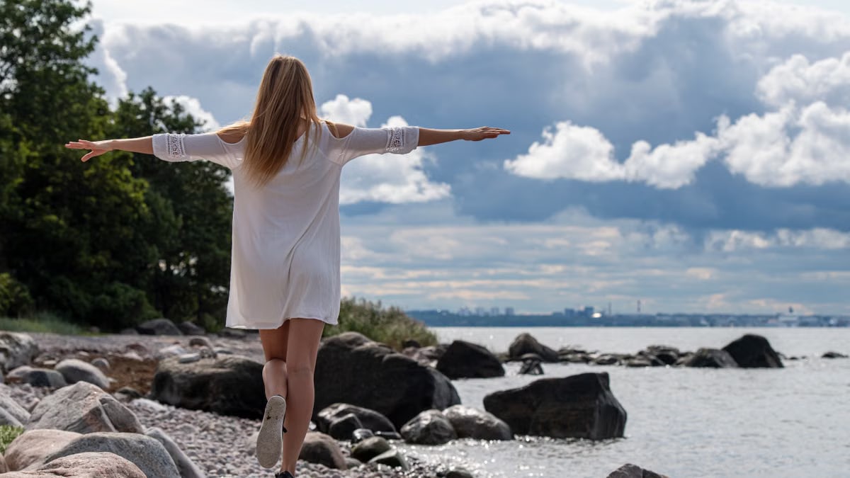A woman in a white dress balances on rocks by the sea in Pringi, Estonia, under a cloudy sky.